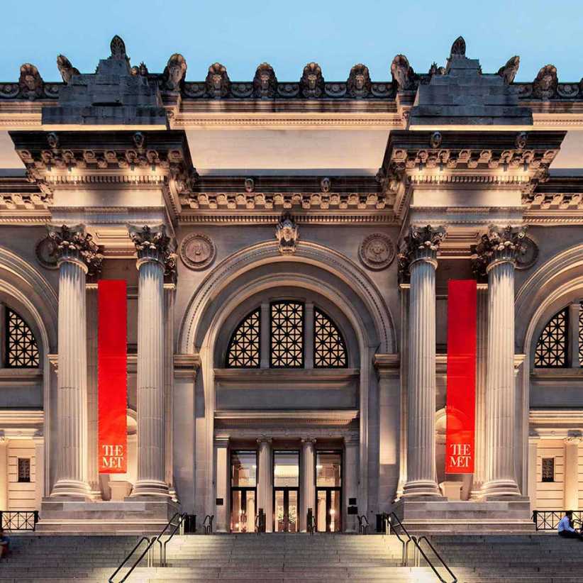 Grand entrance of The Metropolitan Museum of Art (The Met) in New York City, with columns, arches, and red banners at dusk.