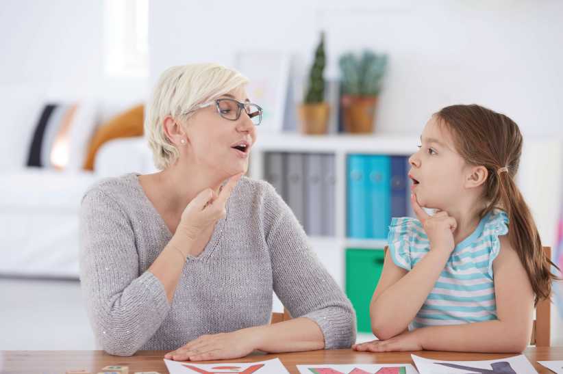 A woman and a young girl sit at a table practicing vocalization techniques together.