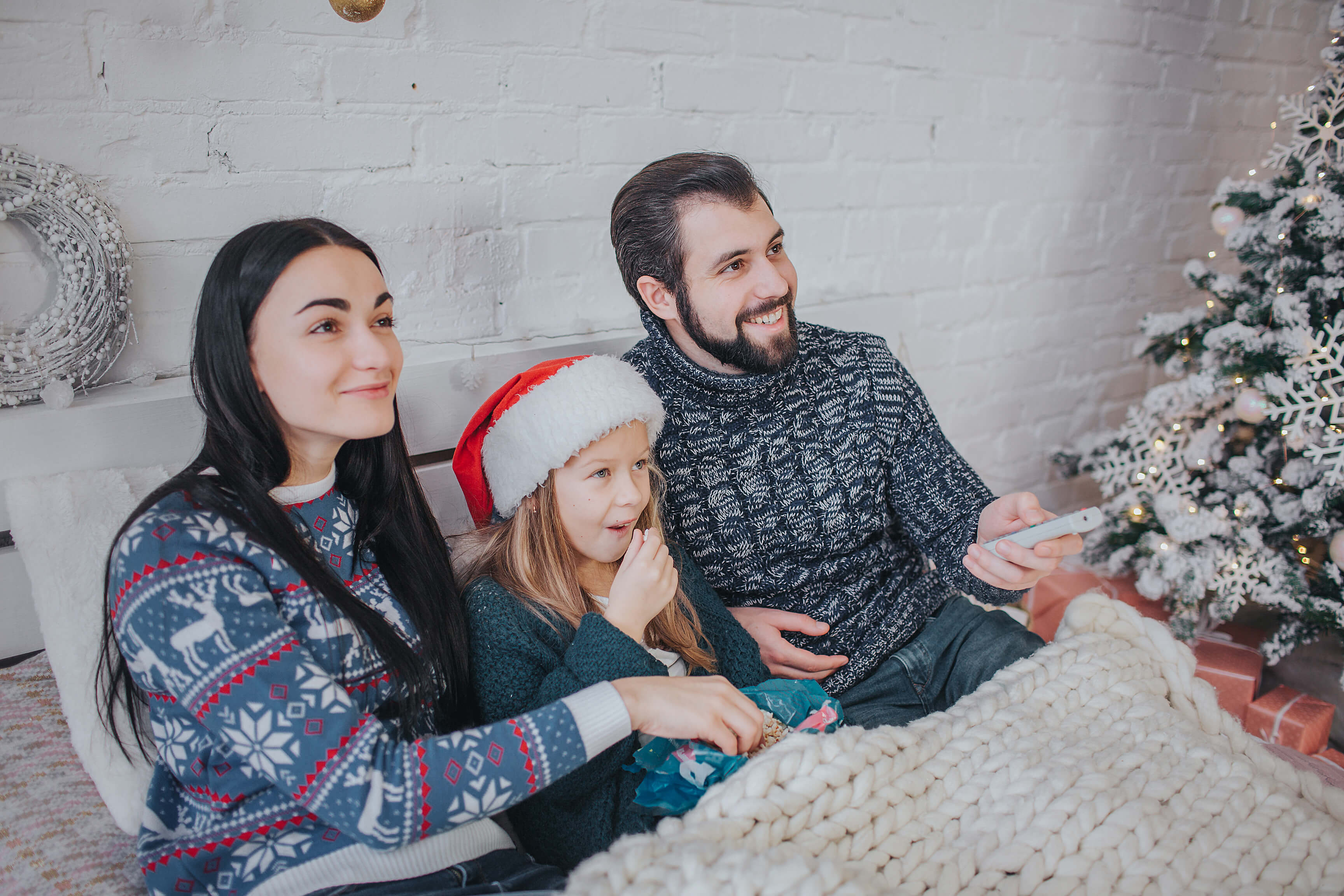 Merry Christmas and Happy New Year . Young family celebrating holiday at home. The Father is holding the remote from the TV. Dad, daughter and mother are watching television.