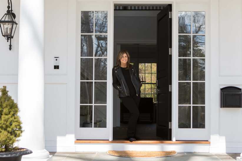 A woman in dark clothing stands smiling in the doorway of a white house with glass-paneled doors and a black mailbox.