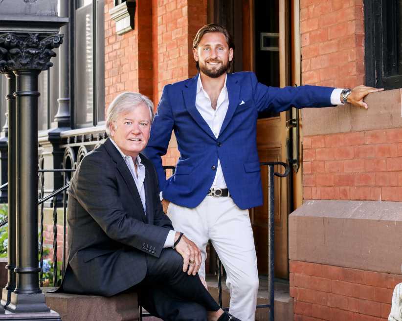 The image shows two men in business attire, one seated and the other standing with an arm resting on a brick wall, outside a building with black railings and a red-brick exterior.