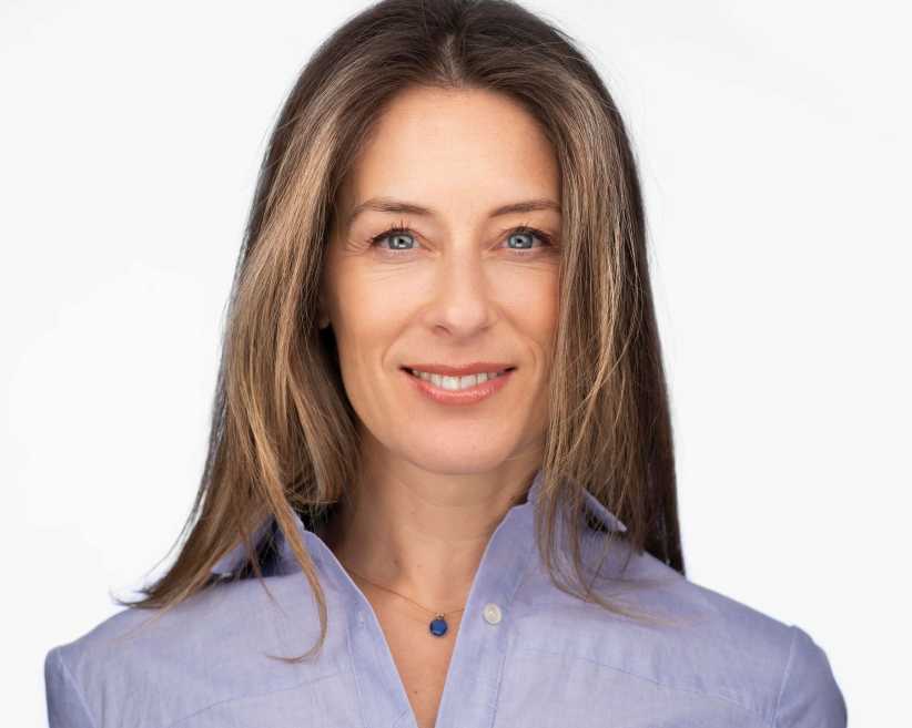 The image shows a woman with long, straight brown hair wearing a light blue shirt and a small pendant necklace, smiling at the camera.