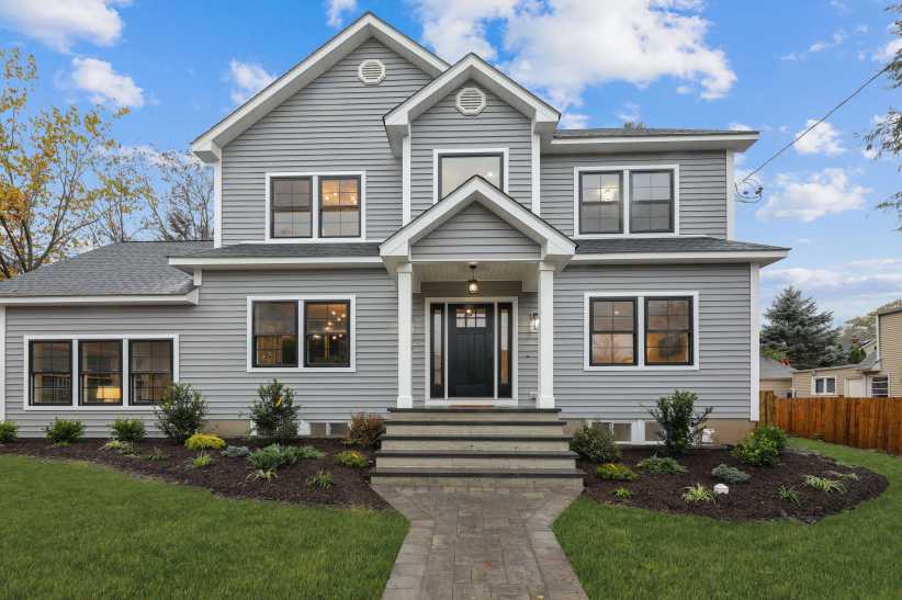 The image shows a modern two-story house with gray siding, black trim around the windows, a prominent front porch with columns, and landscaped front yard under a blue sky.