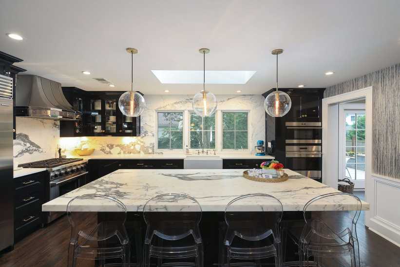 A modern kitchen features a large marble island, clear chairs, dark cabinets, hanging globe lights, and stainless steel appliances .A modern kitchen with a marble island, pendant lights, dark cabinets, glass chairs, and stainless steel appliances under a skylight.