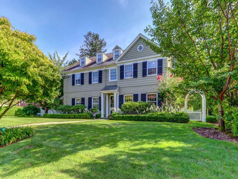 The image shows a large two-story house with beige siding, black shutters, white trim, and a manicured green lawn surrounded by trees and bushes.