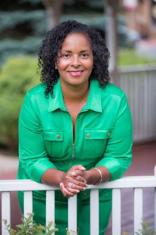 A woman with curly hair in a bright green dress leans on a white fence, smiling outdoors with greenery in the background.