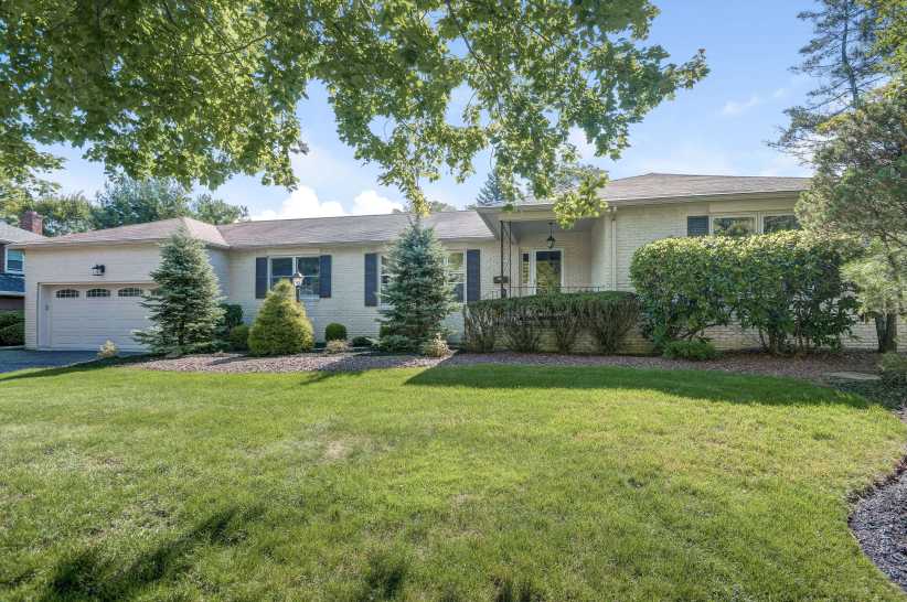 A single-story house with white brick exterior, attached garage, manicured shrubs, and a large green lawn sits under leafy trees.