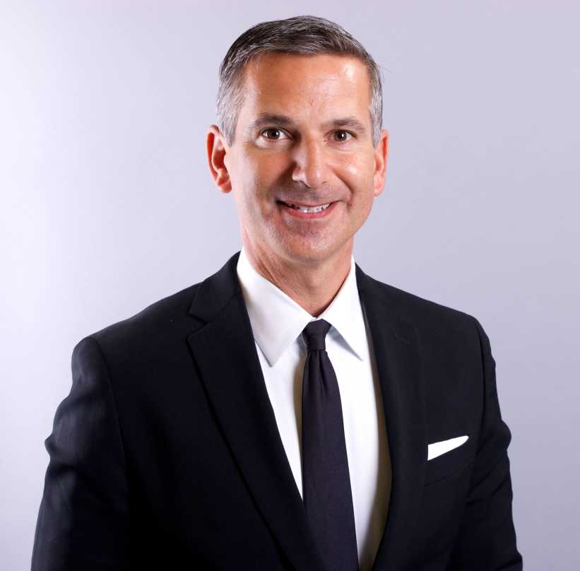 A man in a dark suit, white shirt, and black tie poses against a plain light gray background, facing forward.
