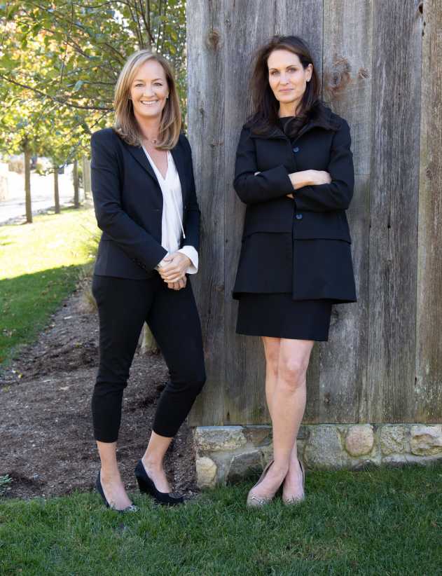 Two women dressed in black business attire stand outdoors on grass, in front of a rustic wooden wall, under leafy trees. Two women dressed in black business attire stand outdoors on grass, in front of a rustic wooden wall, under leafy trees.