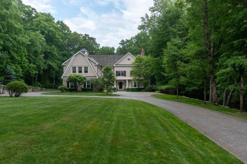 
A two-story house with a circular driveway, surrounded by tall green trees, lush lawns, and a partly cloudy sky overhead.