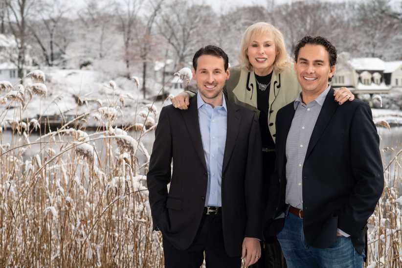 Three people, two men in suits and a woman standing between them, pose outdoors in a snowy landscape near a lake and houses.