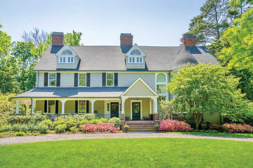 A large colonial-style house features tall chimneys, dormer windows, wraparound porch, and is surrounded by gardens and green lawn.