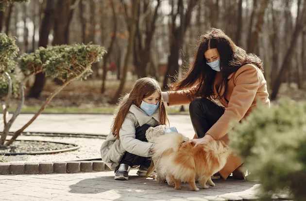 Woman and young girl in coats pet a small fluffy dog on a paved path in a park with trees and trimmed bushes in the background.