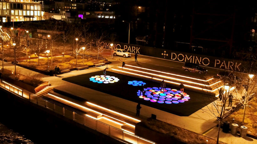 An aerial view of Domino Park's art instillation, Reflect. It features three circles of glowing, multicolored dots with people running around on them to light them up. They glow especially bright in the nighttime of this picture.