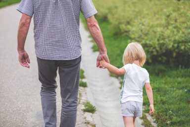 Back view of a man and a small child holding hands while walking down a paved path next to a green field.