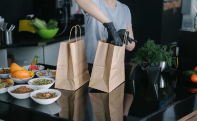A restaurant worker packing up some New York takeout comfort food into paper bags. They're wearing gloves, assumedly for covid safety measures.