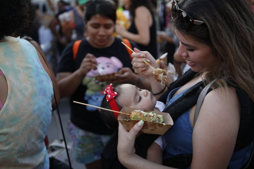 A woman holding a baby close to her bosom. She holds up indecipherable food on a stick for the baby to eat. It's cuter than it sounds.