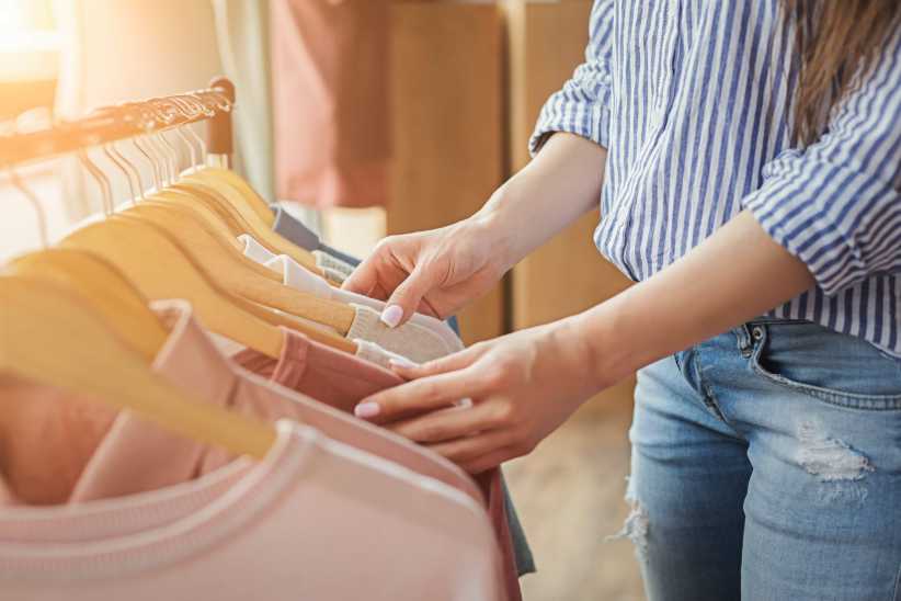 It shows a person wearing a striped shirt browsing clothes on hangers in a store, touching pastel and neutral sweaters.