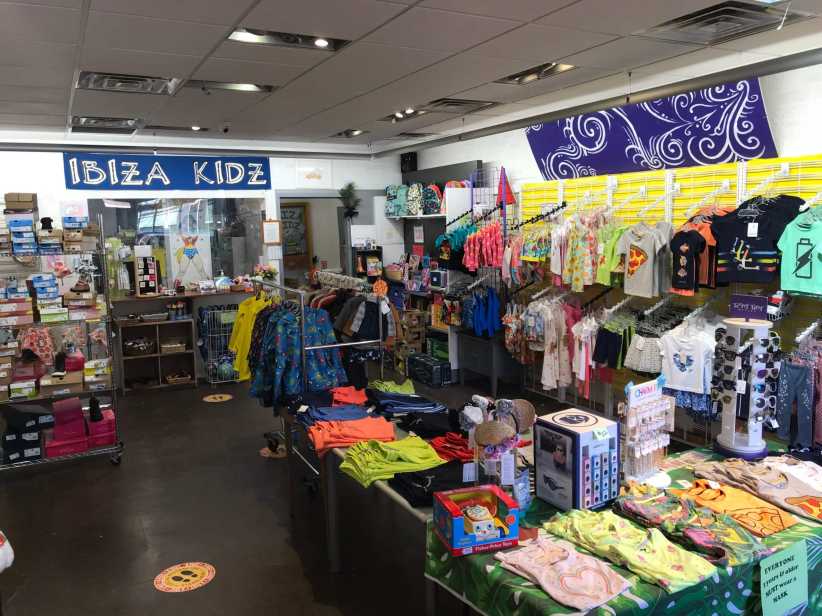 It shows the interior of Ibiza Kidz, a colorful children's boutique filled with racks of bright clothing, shoes, accessories, and toys arranged on shelves and tables.