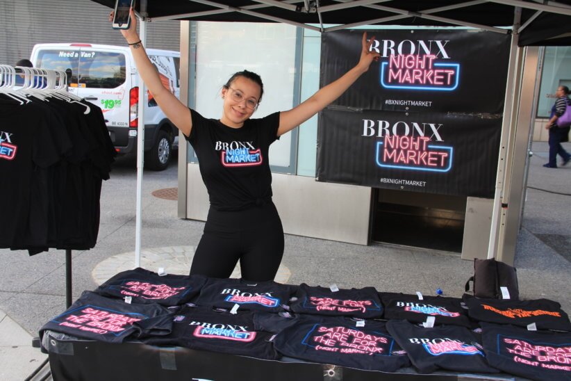 A woman working for the Bronx Night Market posing at a booth displaying Bronx Night Market t-shirts. She poses with both hands up in the air, silly and excited.