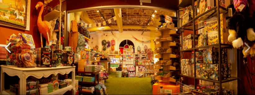 It shows a warmly lit toy store filled with shelves and tables displaying board games, plush toys, puzzles, books, and various colorful children's items.