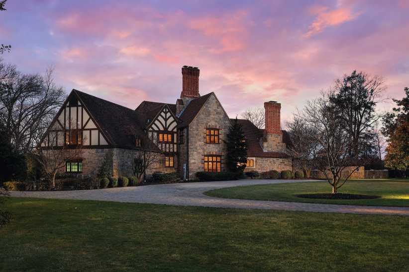 Elegant brick colonial-style house with steep rooflines, dormer windows and lush front lawn lined by mature trees, exuding classic suburban charm.