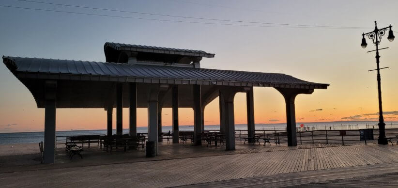Open-air pavilion with metal roof and benches on wooden boardwalk, silhouetted against pastel sunrise sky over calm ocean.