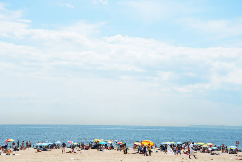 Crowded sandy beach lined with colorful umbrellas and sunbathers beside calm blue ocean under wide, bright, partly cloudy summer sky.