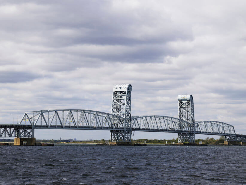Steel lift bridge spanning choppy water under a cloudy sky, with two tall vertical towers and low shoreline visible in the distance.