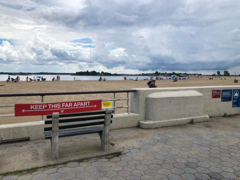 Bench facing sandy beach and calm bay, with red “KEEP THIS FAR APART” distancing sign on railing under cloudy sky.
