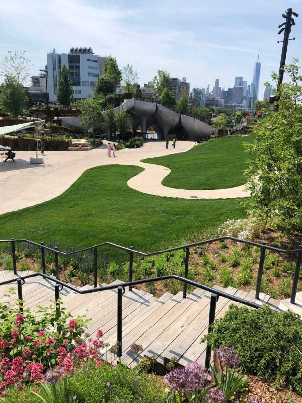 A landscaped park with green lawns, winding paths, stairs, blooming flowers, trees, and city skyline in the background.