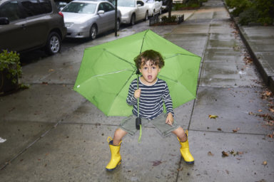 Little boy with a green umbrella in the rain standing on a city sidewalk with yellow rain boots on. He is looking at the camera and making a funny face and his legs are spread apart. Full length shot.