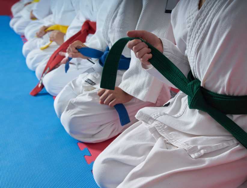 Martial arts students in white uniforms kneel on a blue mat, each holding a colored belt representing their rank, including green, blue, and red.
