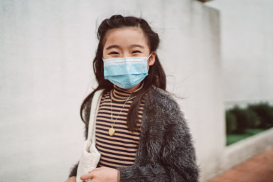 Little girl with medical face mask smiling joyfully at camera in street