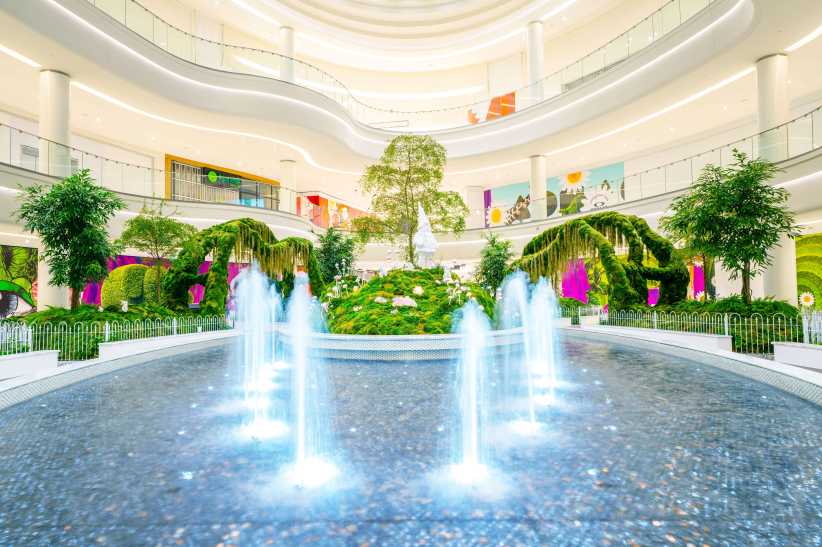 
A modern indoor space features a circular fountain with multiple water jets, lush greenery, and bright, curved architectural balconies.