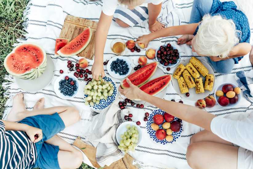 A picnic scene with people sitting on a blanket, sharing watermelon, grapes, blueberries, cherries, peaches, and grilled corn together.