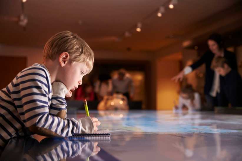 Young boy in striped shirt leans on a lit table, thoughtfully writing in a notebook with a pencil in a dim, museum-like room.