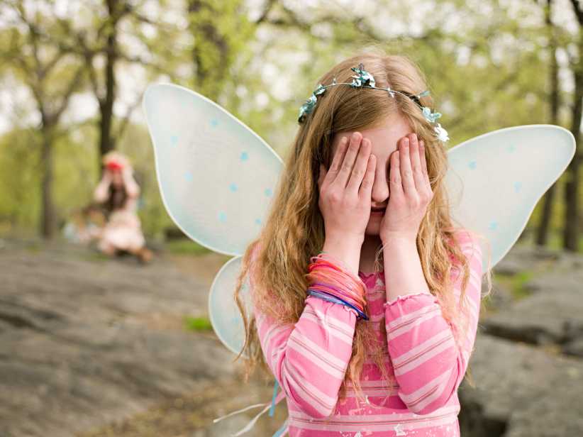 A young girl in a pink outfit with fairy wings and a flower crown covers her face with her hands outdoors, with trees and rocks in the background.