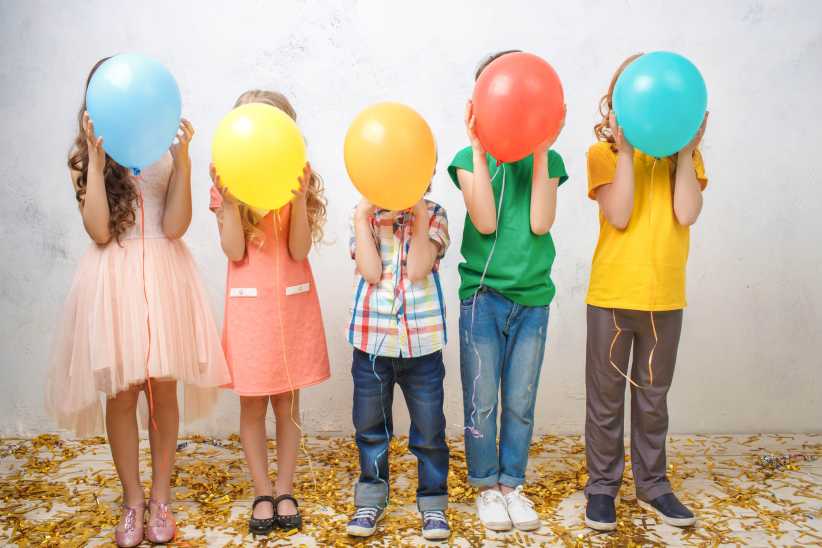 Five children stand in a row, each holding a colorful balloon in front of their face, dressed in bright clothes and surrounded by confetti on the floor.