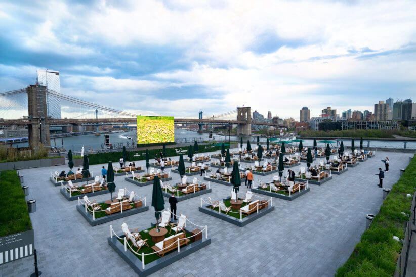 Outdoor rooftop venue with private seating pods, green umbrellas, and a large screen, overlooking the river with bridges and cityscape in the background.