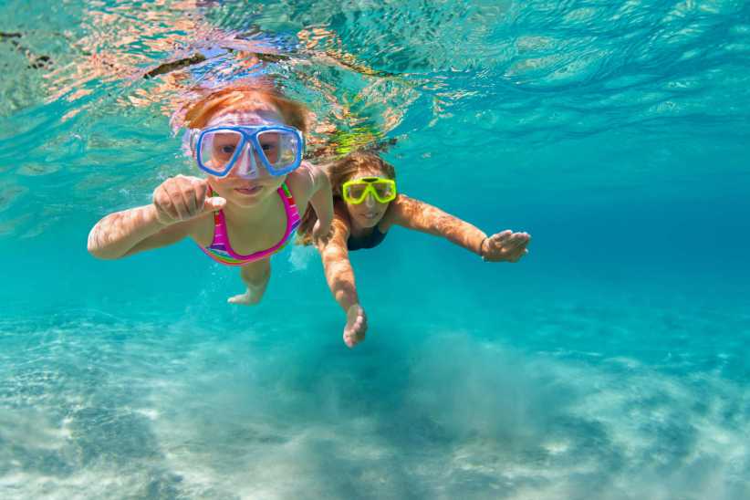 Two children swim underwater in clear turquoise ocean, wearing swim goggles and bright bathing suits, exploring and playing together.