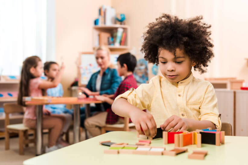 Selective focus of african american kid playing educational game with teacher and children at background in montessori school