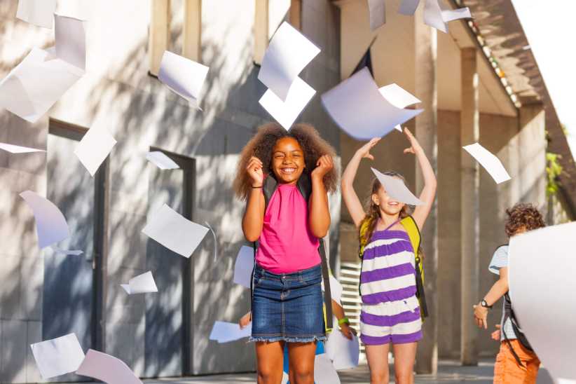 Children celebrate outdoors as papers fly in the air, showing excitement and movement in a sunny urban setting.