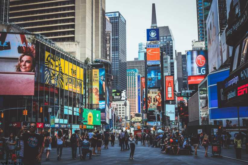 Busy city street in Times Square with crowds walking among tall buildings covered in colorful digital billboards and advertisements.