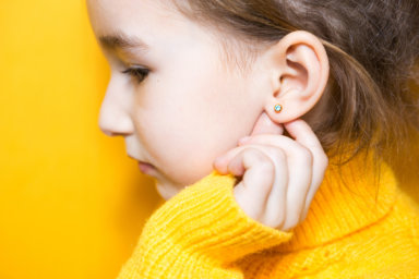 Ear piercing in a child – a girl shows an earring in her ear made of a medical alloy. Yellow background, portrait of a girl in profile.