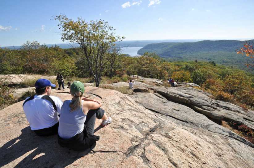 Hikers sit on a rocky ledge overlooking a scenic valley and river at Bear Mountain on a clear, sunny day.