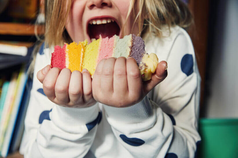 A child in a polka dot shirt excitedly prepares to eat a vibrant slice of rainbow cake, holding it with both hands in front of their mouth.