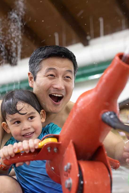 A child and adult in swimwear play together with a red water feature indoors, as water splashes around them in a pool area.