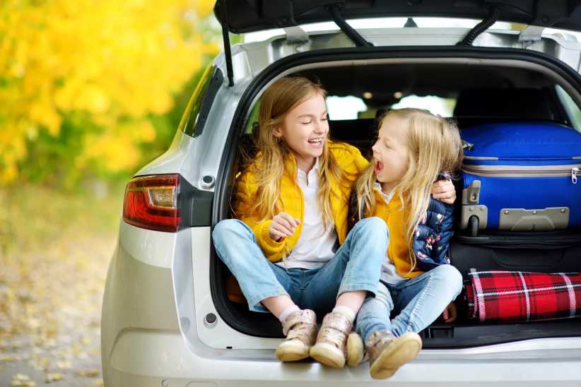Two children sit in the open trunk of a car packed with luggage and blankets, ready for a road trip on a sunny autumn day.