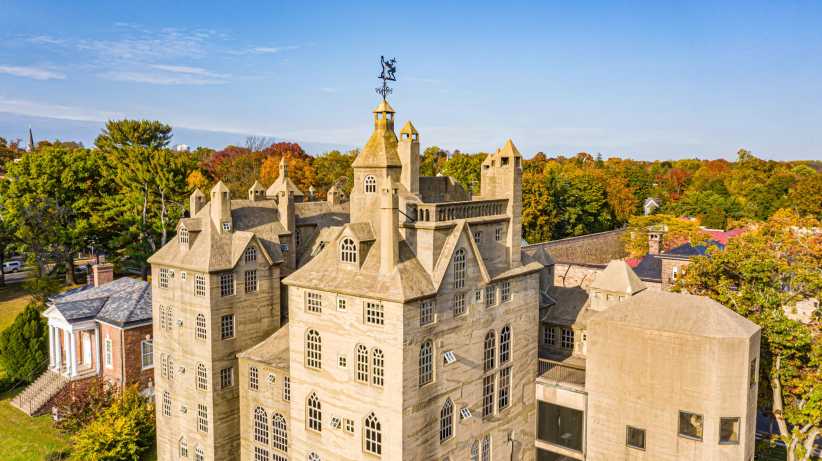 A stone castle-like building with turrets and arched windows stands amid autumn trees, highlighted by a blue sky at the Mercer Museum.